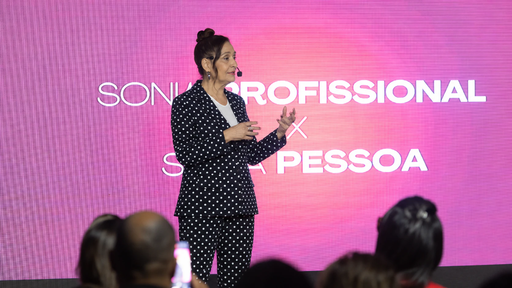 Sonia Abrão no palco, de microfone, falando para a plateia diante de um telão.